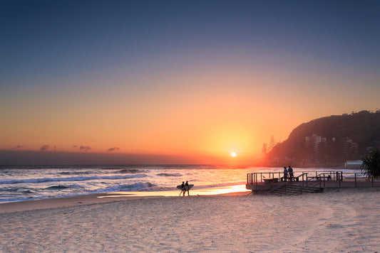 the sun setting across a beach with two surfers walking on the sand